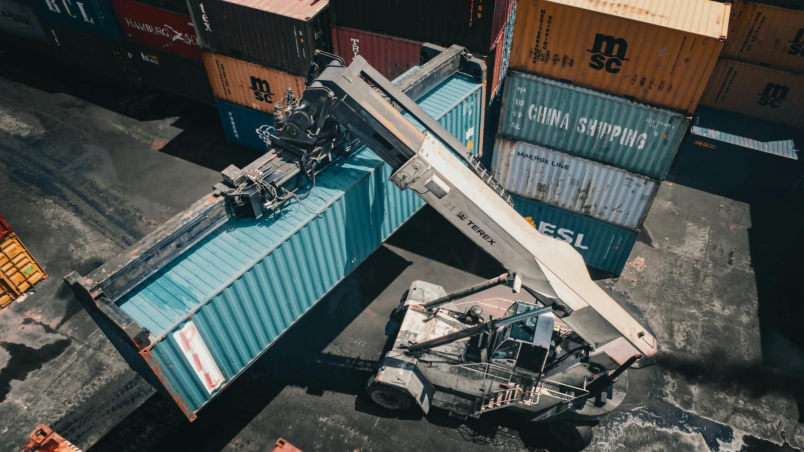 Aerial top-down view of a reach-stacker moving a teal shipping container in a busy port container yard - illustrating the kind of high-value, non-powered asset that GotYou Telematics tracks.